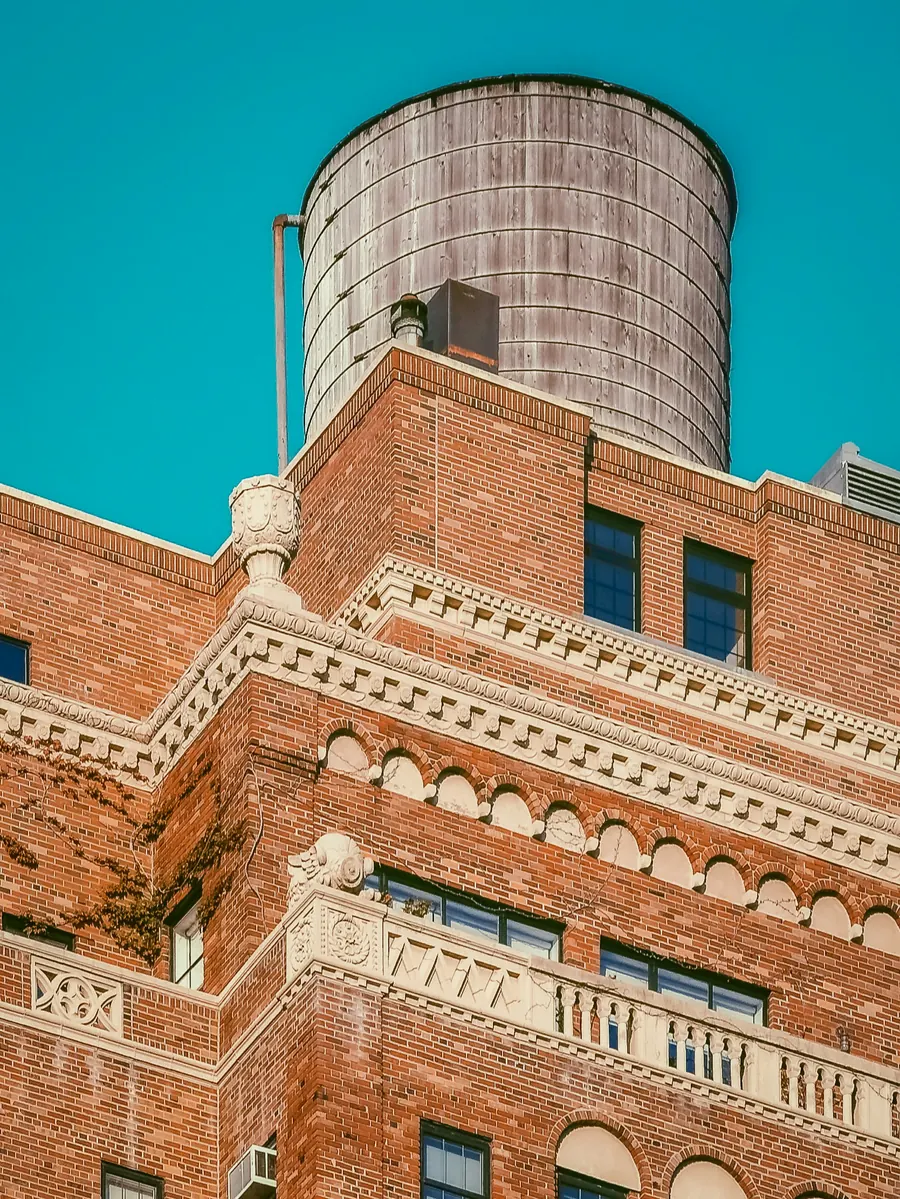 A red-brick pre-war building with a carved cornice and a wooden water tank on the roof against a teal sky
