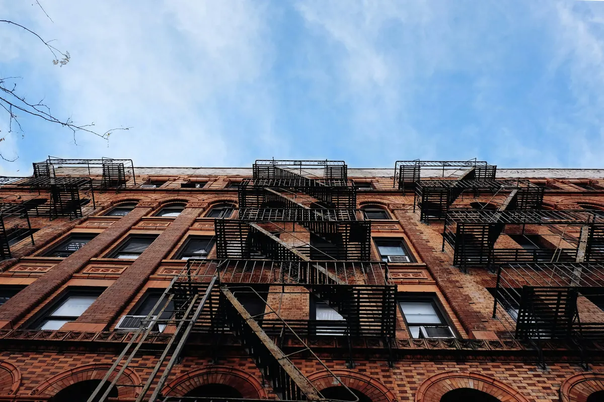 Looking up the red-brick face of a tenement building covered in black fire escapes under a pale blue sky